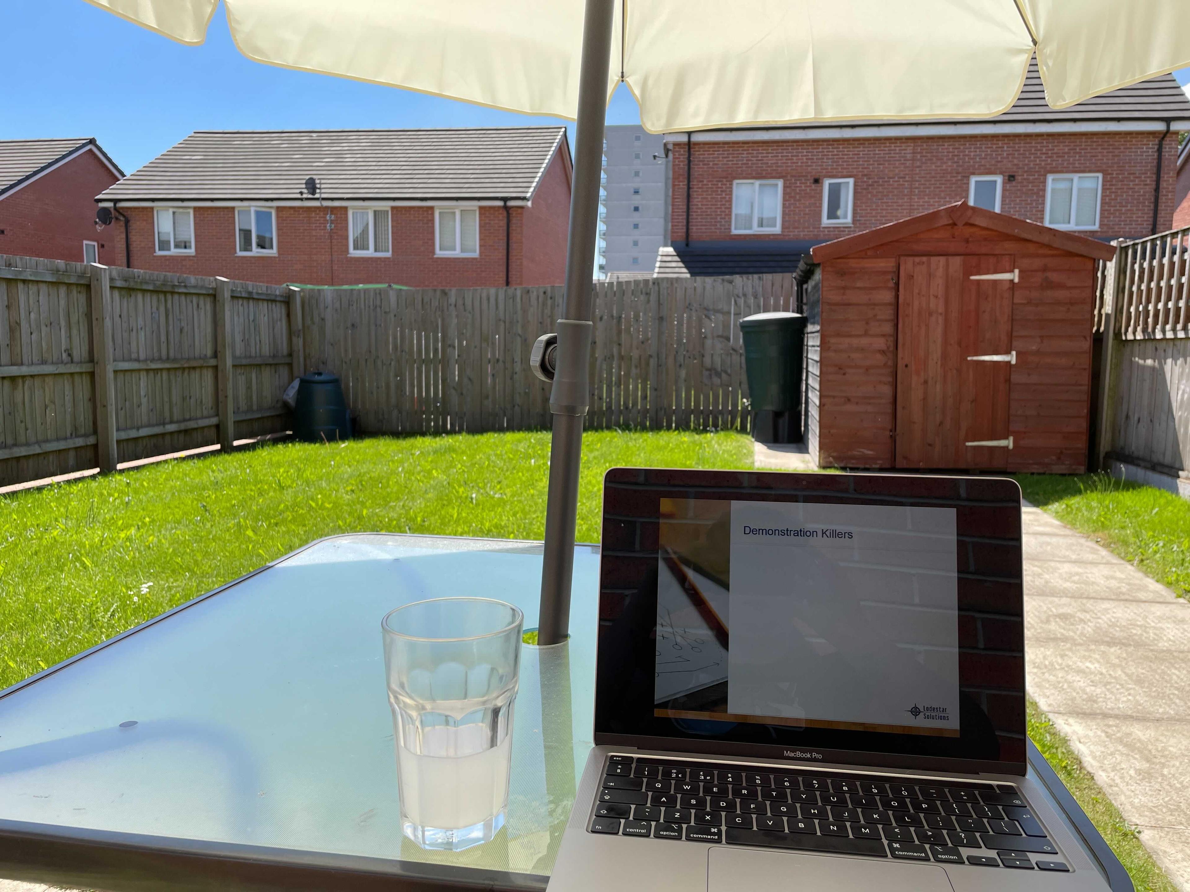 A Macbook pro on a glass garden table, under a cream coloured parasol in a sunny garden